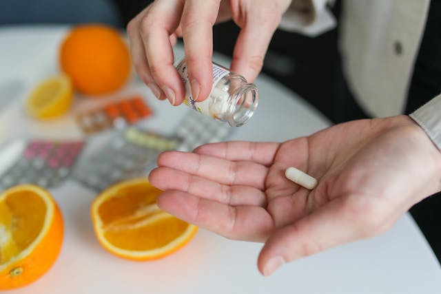 Kézben vitamin kapszula, mellette citrusfélék és gyógyszerek.